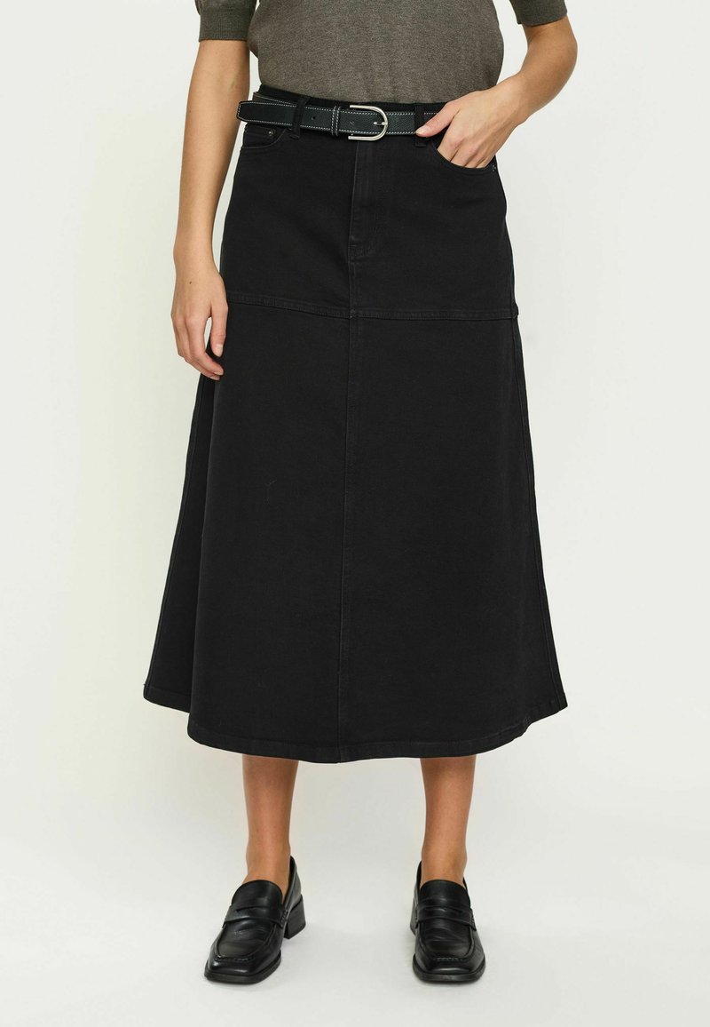 Personne portant un haut gris foncé, une jupe midi noire en A avec ceinture et des mocassins noirs, debout devant un fond clair uni.
