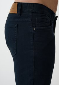 Ciemnoniebieskie dżinsy o slim fit, z tylnymi kieszeniami i brązową skórzaną łatą na pasku. Gładka faktura, zapięcie na jeden guzik.