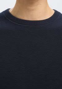 Detailný záber na krk a hornú časť hrudníka osoby, ktorá má na sebe tmavomodrý textúrovaný pletený sveter s okrúhlym výstrihom.