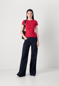 Une jeune femme se tient avec confiance dans un haut rouge ajusté et un pantalon large en jean foncé, tenant un sac à bandoulière noir, devant un fond uni.