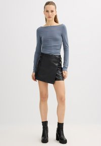 Top de manga larga color azul claro con escote barco, combinado con una falda envolvente de cuero sintético negro con hebillas laterales y botas negras hasta el tobillo.