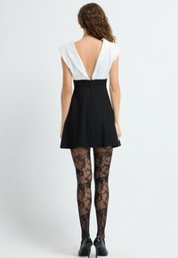 Zwarte skaterjurk met een diepe V-rug en witte kapmouwen, gecombineerd met zwarte bloemenkant panty’s en zwarte hoge hakken.