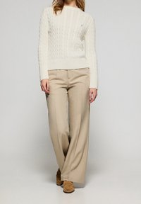 Femme portant un pull en maille torsadée crème, un pantalon beige à jambes larges et des mocassins en daim marron, debout devant un fond clair uni.