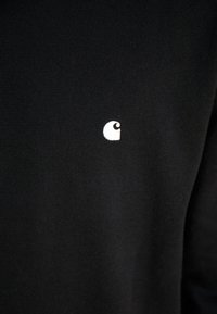 Fekete kötött pulóver, amelyen egy kis fehér logó található a bal mellen. A design egyszerű, sima textúrájú, további minták nélkül.