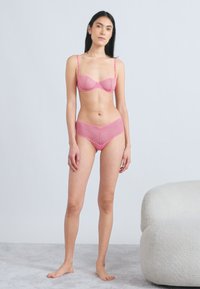 Rožinės nėriniuotos liemenėlės ir atitinkančių kelnaičių rinkinys su geometrine rašto ir reguliuojamomis petnešėlėmis. Minkšta tekstūra, su lankeliais, puošti dekoratyviniais akcentais.