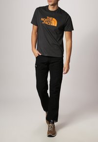 T-shirt gris à manches courtes avec un logo orange, assorti à un pantalon noir avec des poches. Chaussures : baskets marron avec une finition texturée.