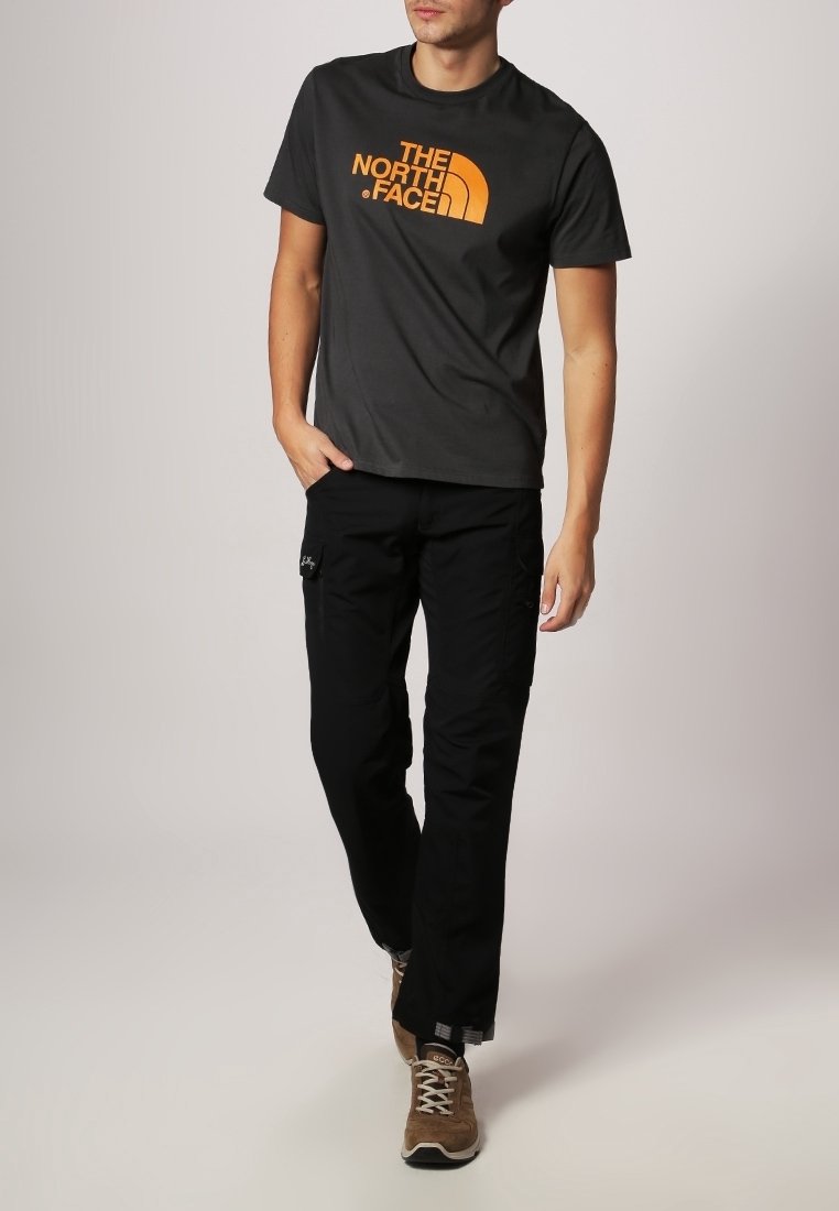 T-shirt gris à manches courtes avec un logo orange, assorti à un pantalon noir avec des poches. Chaussures : baskets marron avec une finition texturée.