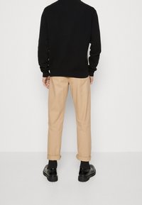 Man i beige upprullade byxor, svart tröja, svarta strumpor och svarta skor som står mot en enfärgad vit bakgrund med ryggen vänd.