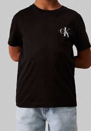 Person iført en sort Calvin Klein Jeans T-shirt med hvidt logo på brystet og lyseblå denimjeans mod en ensfarvet grå baggrund.
