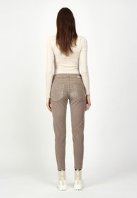 Fitted beige pants with textured back pockets, slim legs, and a waistband detail. Worn with a long-sleeve off-white top and shoes with straps.