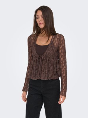 Femme portant un chemisier en dentelle marron à manches longues par-dessus un débardeur marron foncé, associé à un pantalon noir taille haute, debout devant un fond uni.
