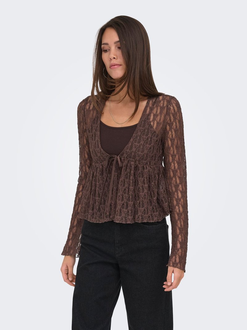 Femme portant un chemisier en dentelle marron à manches longues par-dessus un débardeur marron foncé, associé à un pantalon noir taille haute, debout devant un fond uni.