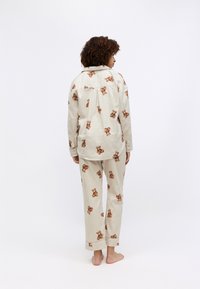 Beige pyjamapakket met een berenprint, bestaande uit een overhemd met knopen en bijpassende broek. Het materiaal ziet er zacht uit met een relaxte pasvorm en zichtbare stiksels.