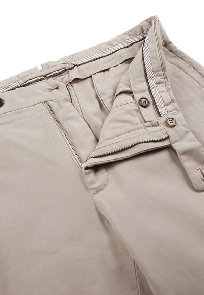 Pantaloni beige con una cerniera aperta, due bottoni in vita e una fodera interna a righe in vita su uno sfondo bianco.