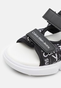 Černé sandály s logem Calvin Klein Jeans, s páskem se vzorem a bílou podrážkou. Zapínání na suchý zip a texturovaná stélka pro pohodlí.