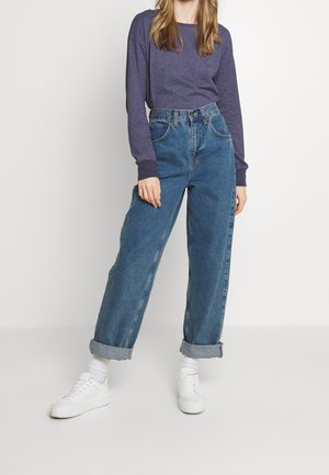 Ciemnofioletowa koszula z długim rękawem, zestawiona z niebieskimi dżinsami z wysokim stanem i podwiniętymi mankietami. Białe buty dopełniają strój.