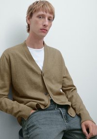 Beige cardigan med V-ringning, ribbade ärmslut och en enkel knäppning. Bärs över en vit t-shirt, matchad med mörka jeans. Mjuk textur.
