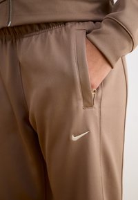 Brūnas Nike sporta bikses ar teksturētu audumu, elastīgu jostasvietu un sānu kabatu. Apakšējā kājā ir maza balta logo.