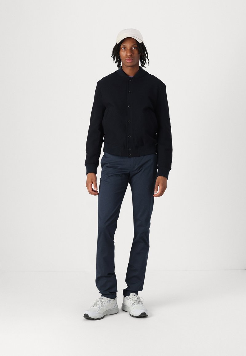 Giacca bomber nera con chiusura a bottoni, pantaloni blu navy e scarpe da ginnastica bianche. Gli accessori includono un cappellino beige chiaro. Sfondo semplice.