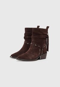 Botines de ante marrón con caña arrugada, detalles de tachuelas plateadas, acentos de borlas y tacones de madera apilados bajos sobre fondo neutro.