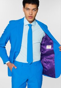 Heled sinine ülikond koos sobiva lipsu, helesinise särgi ja jakis nähtava lilla voodriga. Sile kangas ja kohandatud disain.