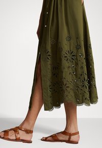 Olivgrön klänning med blommönster i utskärning längs fållen, med en sidoklyvning. Åtföljs av bruna platta sandaler med spänne.