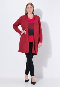 Rode lichtgewicht cardigan met een ontspannen pasvorm, open voorkant en twee voorzakken, gedragen over een kleurrijke gradient top en zwarte legging.