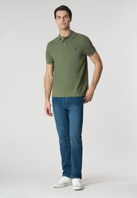 Polo shirt verde oliva con colletto a strisce bianche e blu, abbinato a jeans blu e sneakers bianche. Taglio e design semplice e classico.