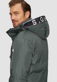 Szara ocieplana kurtka z czarną i białą kołnierzem z logo, zamkiem błyskawicznym w bocznej kieszeni i kapturem, widoczna z tyłu.