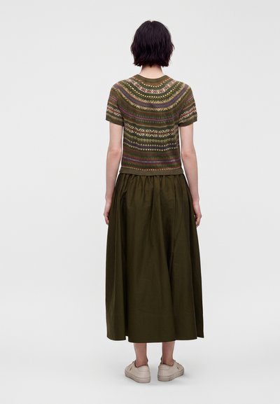 Femme aux cheveux courts et foncés portant un pull à manches courtes à motifs et une longue jupe vert olive, debout de dos.