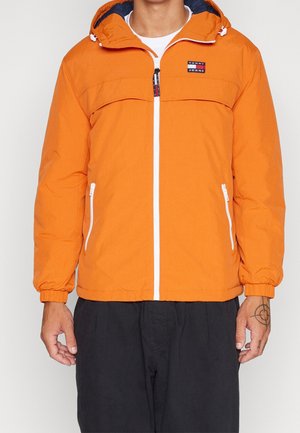 Mann trägt eine hell orange Kapuzenjacke mit weißem Reißverschluss und Taschen, kombiniert mit schwarzen Hosen, steht vor einem einfarbigen Hintergrund.