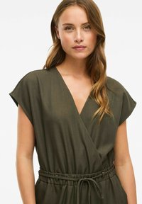 Joven mujer con cabello castaño claro y ondulado, que lleva un vestido envolvente sin mangas de color verde oliva con un cinturón atado en la cintura.