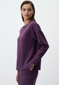 Felpa viola con scollo rotondo e maniche lunghe, caratterizzata da una vestibilità rilassata e fessure laterali. Il materiale appare morbido e dalla texture interessante.