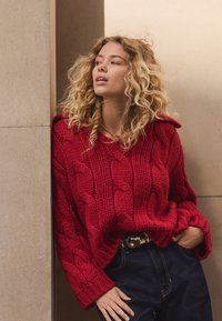 Red knitted cable sweater with wide sleeves, worn over dark denim jeans. Textured fabric with a loose fit and a ribbed collar detail.