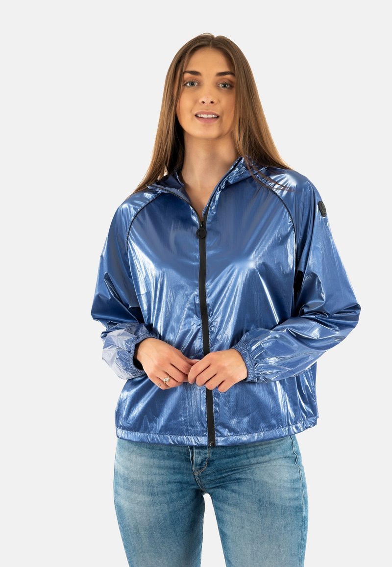 Blauwe reflecterende windbreaker jacket met een rits aan de voorkant, zwarte accenten, elastische manchetten en een staande kraag. Draagt over denim.