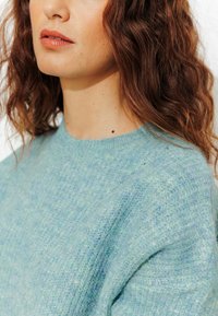 Pull en tricot bleu clair avec une finition douce et texturée. Présente un col rond et une coupe légèrement décontractée, mettant en valeur le détail du motif en tricot.