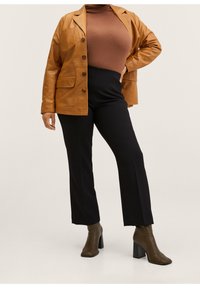 Personne portant une veste en cuir marron, un col roulé marron, un pantalon noir et des bottines marron à talons, debout avec une main sur la hanche.