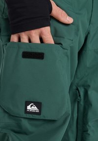 Mano con manica nera inserita nella tasca di pantaloni Quiksilver verde scuro, dotata di patta in Velcro.