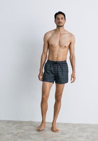 Pantaloni de baie negri cu un model repetitiv cu logo-ul "HUGO", dotati cu un waistband elastic si snur ajustabil. Modelul sta desculț.