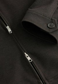 Mörkgrå jacka med en slät textur, metalldragkedja och en knappdetalj vid ärmslutet. Designen är strukturerad med krage.