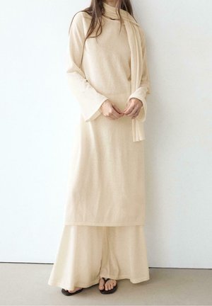 Femme portant une tunique crème à manches longues, pantalon large assorti, une écharpe et des sandales noires, debout contre un mur blanc.