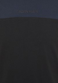 Camiseta de algodón negra con panel superior azul oscuro. Presenta un sutil logo de Calvin Klein en un color a juego en el pecho. Textura suave.