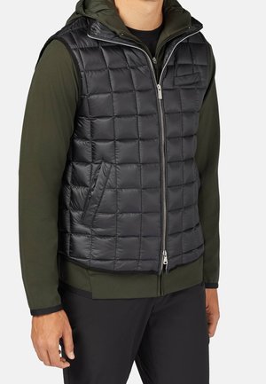 Uomo che indossa un gilet nero trapuntato senza maniche sopra una felpa con cappuccio verde oliva a maniche lunghe e pantaloni neri, in piedi su sfondo bianco.