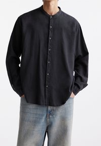 Zwarte button-up shirt met een hoge kraag, lange mouwen en een relaxte pasvorm, gecombineerd met lichtblauwe denim jeans. Zachte stoftextuur.