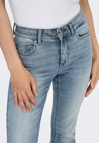 Gros plan d'une personne portant un jean bleu clair délavé et un haut blanc, avec une main posée sur la cuisse.