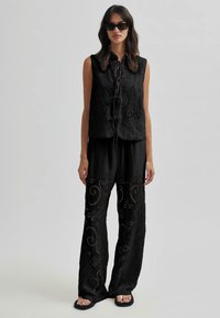 Top negru brodat fără mâneci, cu un nod în față și pantaloni largi asortați, cu modele intricate în spirale și detalii decupate.