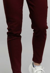 Pantalons ajustés bordeaux en tissu extensible, présentant une texture lisse et un design fuselé, associés à des baskets blanches.