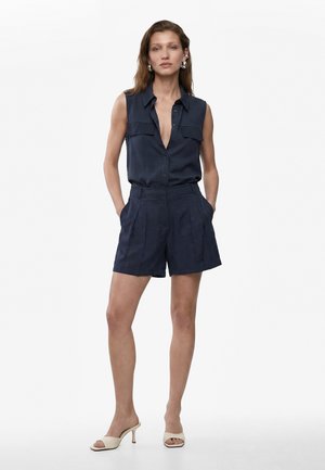 Kvinde står med hænder i lommerne iført ærmeløs marineblå knapbluse, matchende shorts med høj talje, sølvøreringe og beige hælede sandaler.