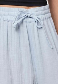 Svetlo modri, teksturirani hlačni iz materiala s fleksibilnim pasom in zavezanim mlinom spredaj, ki prikazujejo sproščen kroj in mehak padec.