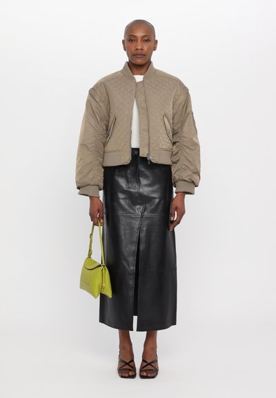 Khatuluh beige bomber jaket, desain cropped, kerah berulir, rok midi kulit hitam, tas tangan hijau, sepatu hak hitam tali.